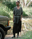 A woman stands outdoors near a pond, wearing the Pleated Skirt with black boots and a grey cable-knit sweater. She has another matching sweater tied at her waist. A green car is on the left, while trees and forest hues fill the background.