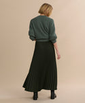 A person with short blonde hair, wearing a green cable knit sweater, Pleated Skirt, and black ankle boots stands facing away against a beige background.