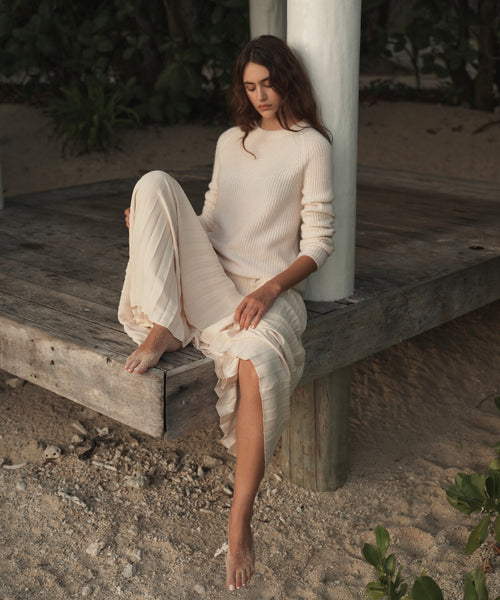 A woman sits barefoot on a weathered wooden platform outdoors, wearing a cream ribbed knit long-sleeve top and matching pleated skirt.