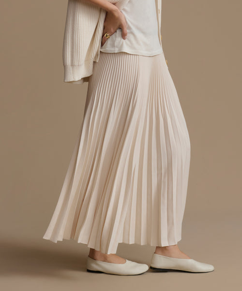 Person wearing an ivory pleated skirt and white top against a brown background