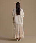 Woman wearing an ivory sweater and pleated skirt against a brown background