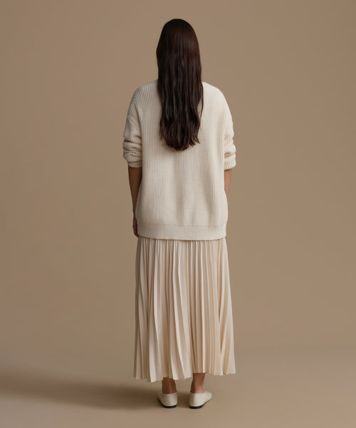 Woman wearing an ivory sweater and pleated skirt against a brown background