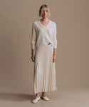 A woman stands against a plain beige studio background wearing a cream V-neck knit sweater, a matching pleated midi skirt, and white flats.