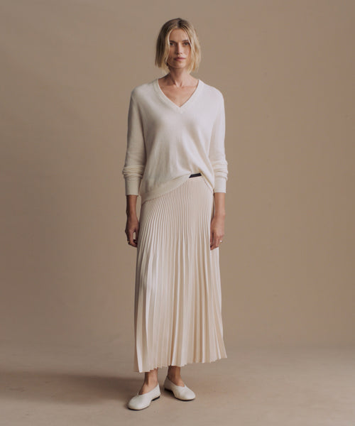 A woman stands against a plain beige studio background wearing a cream V-neck knit sweater, a matching pleated midi skirt, and white flats.