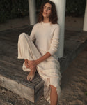 A woman wearing a cream ribbed knit long-sleeve top and matching pleated skirt sits barefoot on a weathered wooden platform outdoors.