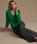 Woman wearing a green sweater and slate pleated skirt sitting on a beige background