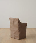 A brown, padded chair with a shiny fabric cover sits on a light wood floor by a white wall. Protect it and keep it looking new with the Outdoor Montecito Dining Arm Chair Cover, designed for a perfect fit and lasting durability.