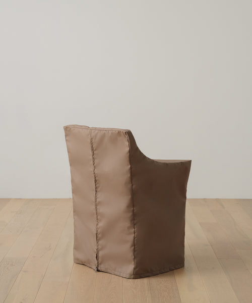 A brown, padded chair with a shiny fabric cover sits on a light wood floor by a white wall. Protect it and keep it looking new with the Outdoor Montecito Dining Arm Chair Cover, designed for a perfect fit and lasting durability.