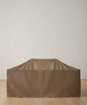 The Outdoor Vista Dining Table Cover, made of durable brown polyester, protects a large rectangular dining table placed on a light wood floor beside a plain white wall.