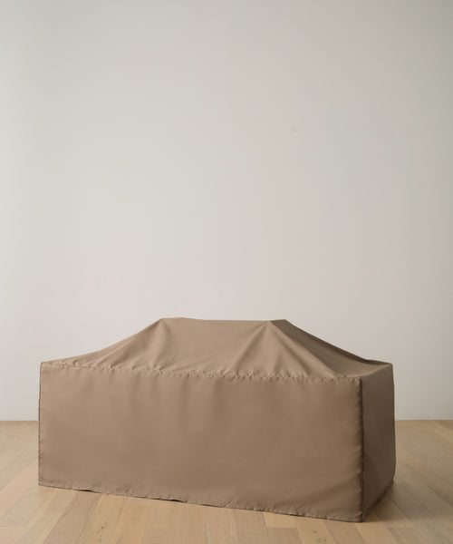 A tan, rectangular object is shielded by the Outdoor Vista Dining Table Cover, made of durable polyester. The fitted cover rests smoothly with gentle creases on a light wooden floor against a plain white wall.