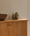 A wooden cabinet with vertical panels displays three Porta Ceramic Vases in muted tones and a rustic stone bowl, bringing timeless style to your home décor against a plain, light-colored wall.
