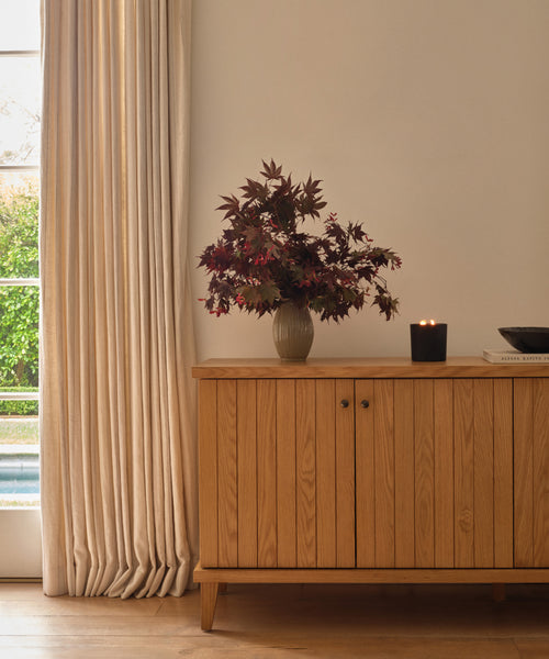 The Porta Ceramic Vase, styled with dark red leaves, stands on a wooden sideboard beside a lit black candle and book, set against cream walls and beige curtains—ideal for adding elegant style to your home décor.