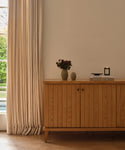A wooden sideboard stands against a cream wall by a window with beige curtains. On it sits the Porta Ceramic Vase filled with flowers, stacked books, and a lit candle—perfect touches for elegant home décor as sunlight filters in.