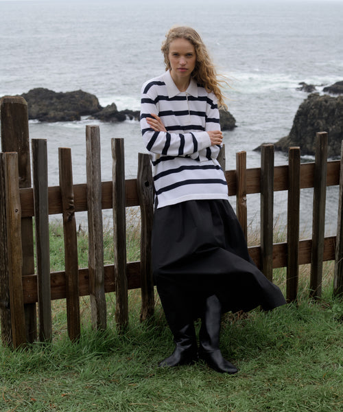Woman in a Navy Stripe post pullover worn by a person with a neutral background and black skirt standing by a wooden fence with ocean in the background