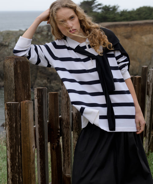 Woman wearing a navy stripe post pullover standing outdoors by a wooden fence.