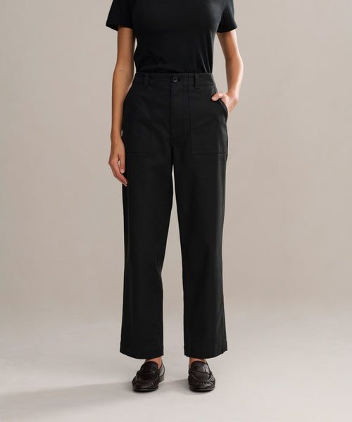 A person stands against a plain light background with one hand in their pocket, wearing a black fitted short-sleeve shirt, Ranger Trouser in black lightweight twill with front pockets, and black shoes.