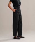A person in black Ranger Trouser, a black short-sleeve shirt, and black loafers stands with hands in pockets against a plain light-colored background.