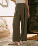 A person wearing Relaxed Trouser with a high waist and wide-leg silhouette stands on a stone floor in sunlight near a glass door, paired with white sandals. Their upper body and face are out of frame.
