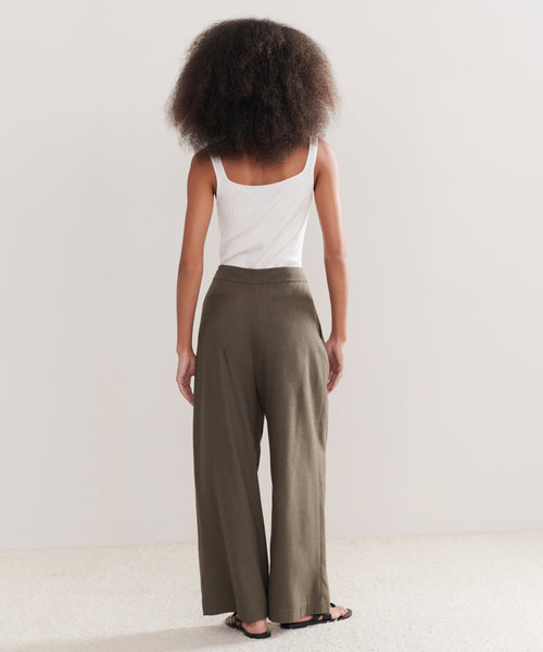 A person with voluminous, curly hair stands facing away, wearing a white sleeveless top, Relaxed Trouser in raw silk olive green, and black sandals against a plain light background.