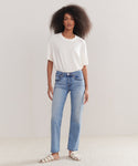 A woman with voluminous curly hair and a neutral expression stands against a plain light background, wearing a loose white t-shirt, Mid Rise Stovepipe jeans in a straight-leg fit, and white sandals.