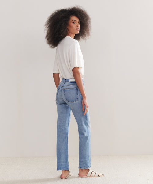 A woman with voluminous curly hair stands sideways, looking over her shoulder. She wears a white t-shirt, Mid Rise Stovepipe jeans, and white sandals against a plain light background.