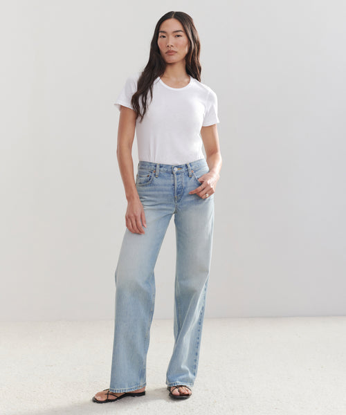 A woman stands against a plain background wearing a white t-shirt, the Mid Rise Wide Leg light blue jeans, and black sandals, with one hand in her pocket.