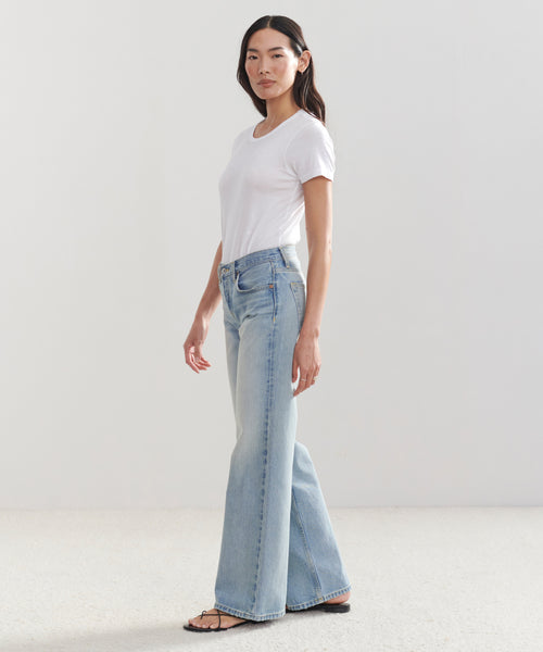 A woman stands in a minimalist room, confidently facing the camera in a white t-shirt, black sandals, and the Mid Rise Wide Leg jeans in light blue stretch denim.