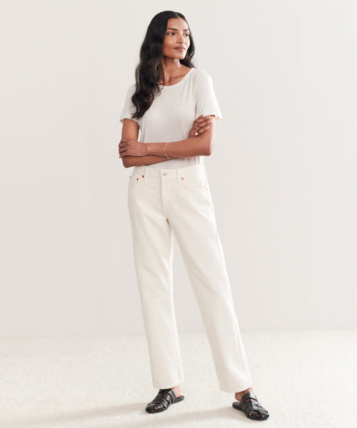 A person stands against a plain background, wearing a light short-sleeve t-shirt, Easy Straight Crop vintage white jeans, and black sandals, with arms crossed and a relaxed expression.