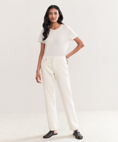 A woman with long wavy hair stands against a plain background, wearing a short-sleeve white t-shirt, Easy Straight Crop vintage white jeans, and black sandals. She has a neutral expression and rests one hand on her hip.