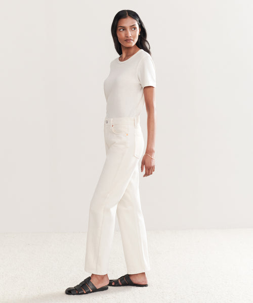 A woman stands sideways in a minimal, light-toned room, wearing a short-sleeved white top and Easy Straight Crop vintage white jeans by Re/Done with black sandals on a textured floor.