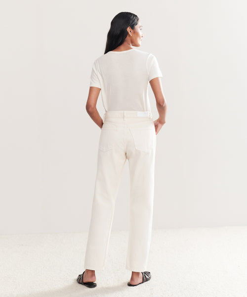 A person with long dark hair, wearing a straight crop white t-shirt, the Easy Straight Crop jeans in white by Re/Done, and black slide sandals stands facing away from the camera against a plain light background.