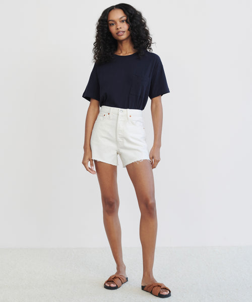 A woman with curly hair stands against a white background, wearing a black short-sleeve t-shirt, Low Slung Short in vintage white, and brown sandals. She has a relaxed posture and a neutral expression.