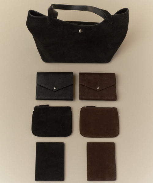A black tote bag is shown at the top, with six small leather accessories—including a Reece Leather Wallet and a brown mid-size wallet—arranged in two vertical columns below it on a beige surface.