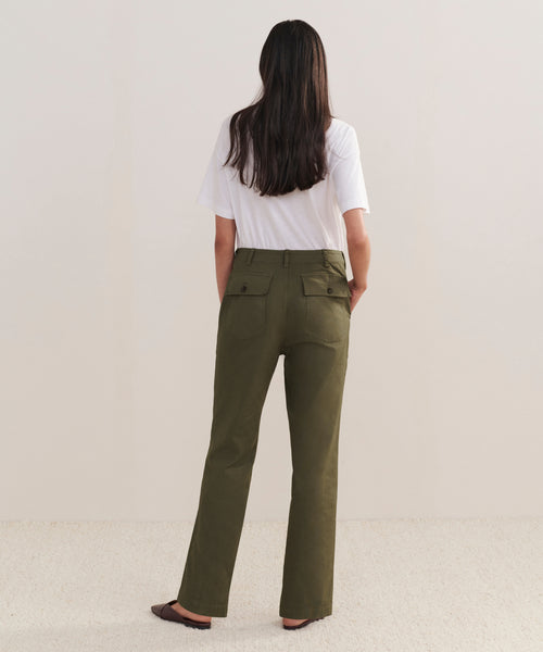 A person with long brown hair, wearing a white t-shirt, Relaxed Army Pant, and brown flats stands facing away from the camera against a plain light background.