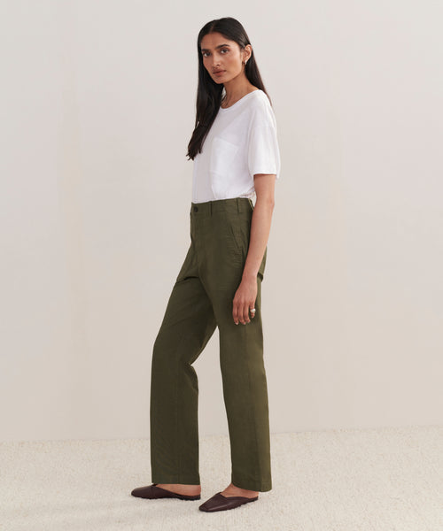 A woman stands against a plain light background, wearing a white t-shirt and the Relaxed Army Pant in olive green. She is sideways, looking at the camera with a neutral expression and wearing dark brown flat shoes.