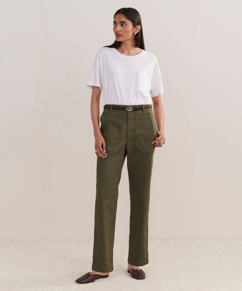A woman stands against a plain background wearing a white t-shirt tucked into high-waisted Relaxed Army Pants with a belt and large pockets, paired with dark brown flat shoes.