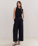 A woman with long, wavy hair stands against a plain background wearing the Relaxed Trouser, a wide-leg silhouette pant, paired with a sleeveless black top and black sandals. Her hands are in her pockets and she appears relaxed.