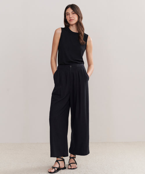 A woman with long, wavy hair stands against a plain background wearing the Relaxed Trouser, a wide-leg silhouette pant, paired with a sleeveless black top and black sandals. Her hands are in her pockets and she appears relaxed.