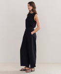 A woman with long brown hair stands against a plain background wearing the Relaxed Trouser in a wide-leg silhouette, paired with a sleeveless black top and black sandals. Her hands are in her pockets.