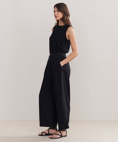 A woman with long brown hair stands against a plain background wearing the Relaxed Trouser in a wide-leg silhouette, paired with a sleeveless black top and black sandals. Her hands are in her pockets.