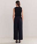 A woman faces away from the camera, wearing Relaxed Trouser in a wide-leg silhouette with a sleeveless black top and black sandals, set against a plain light background.