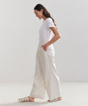 A woman with long dark hair, wearing a white t-shirt, Relaxed Trousers, and light sandals stands in profile with hands in her pockets, smiling against a plain light background.