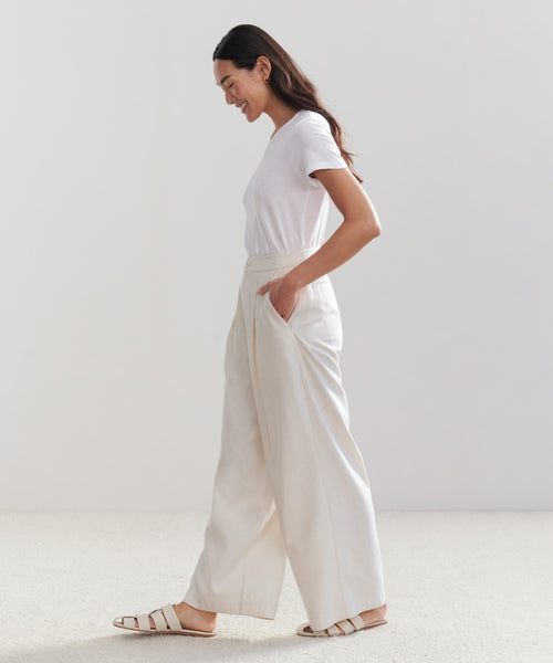 A woman with long dark hair, wearing a white t-shirt, Relaxed Trousers, and light sandals stands in profile with hands in her pockets, smiling against a plain light background.