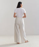 A woman stands barefoot on a light carpet, facing away, wearing the Relaxed Trouser—high-waisted with wide legs—and a white T-shirt. Her dark hair is loose down her back against a plain, light background.
