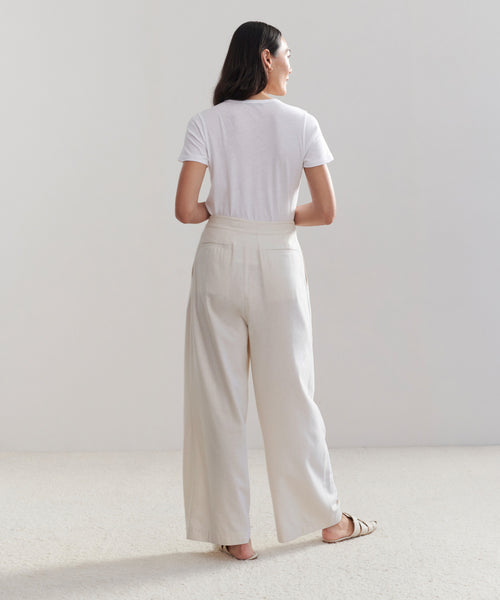 A woman stands barefoot on a light carpet, facing away, wearing the Relaxed Trouser—high-waisted with wide legs—and a white T-shirt. Her dark hair is loose down her back against a plain, light background.