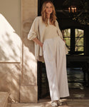 A woman with long, wavy hair stands in a sunlit doorway, wearing Relaxed Trouser paired with a beige button-up shirt, tank top, and white sandals. Her laid-back posture is accentuated by one hand in her pocket.