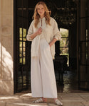 A woman with long blonde hair stands in a doorway, confidently wearing relaxed wide-leg trousers (Relaxed Trouser), a loose cream shirt over a polo top, and white sandals, with one hand in her pocket.