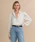 A woman with a short blonde bob wears the cream-colored satin Remy Shirt with puffed sleeves, tucked into high-waisted blue jeans and finished with a black belt, standing against a beige background with a neutral expression.