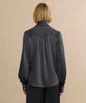 A person with blonde, chin-length hair is seen from behind wearing the Remy Shirt, featuring vintage-inspired satin fabric, puffed sleeves, and buttoned cuffs, paired with black pants against a plain beige background.