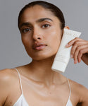 A woman with smooth, glowing skin holds a tube of Renew Face Polish near her face, featuring upcycled pineapple exfoliants. She wears a white spaghetti strap top and has a neutral expression against a plain gray background.